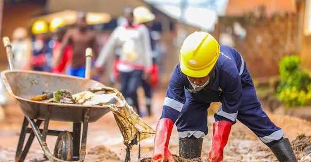 A volunteer in protective gloves and helmet clearing mud and debris from a blocked drainage channel